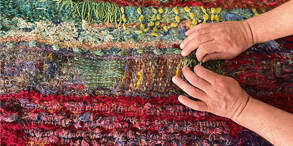 Mãos tocando uma tapeçaria colorida feita à mão, simbolizando o prazer de criar com liberdade, sensibilidade e expressão no bordado artesanal.