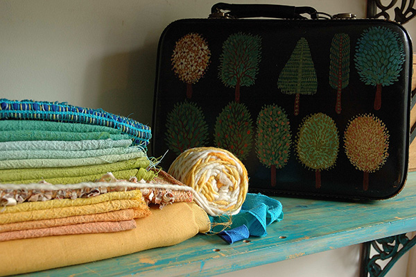 Retalhos para aplicação em bordados. Uma série de retalhos de tecido, todos de tom terossos, empilhados uns sobre os outros. Ao fundo da cena, uma maleta de coura com árvores pintadas.