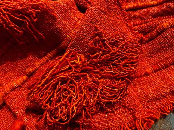 Close de uma obra têxtil em tons intensos de vermelho, com diferentes tramas e relevos bordados que se sobrepõem, formando uma superfície densa de fios, texturas e entrelaçamentos.
